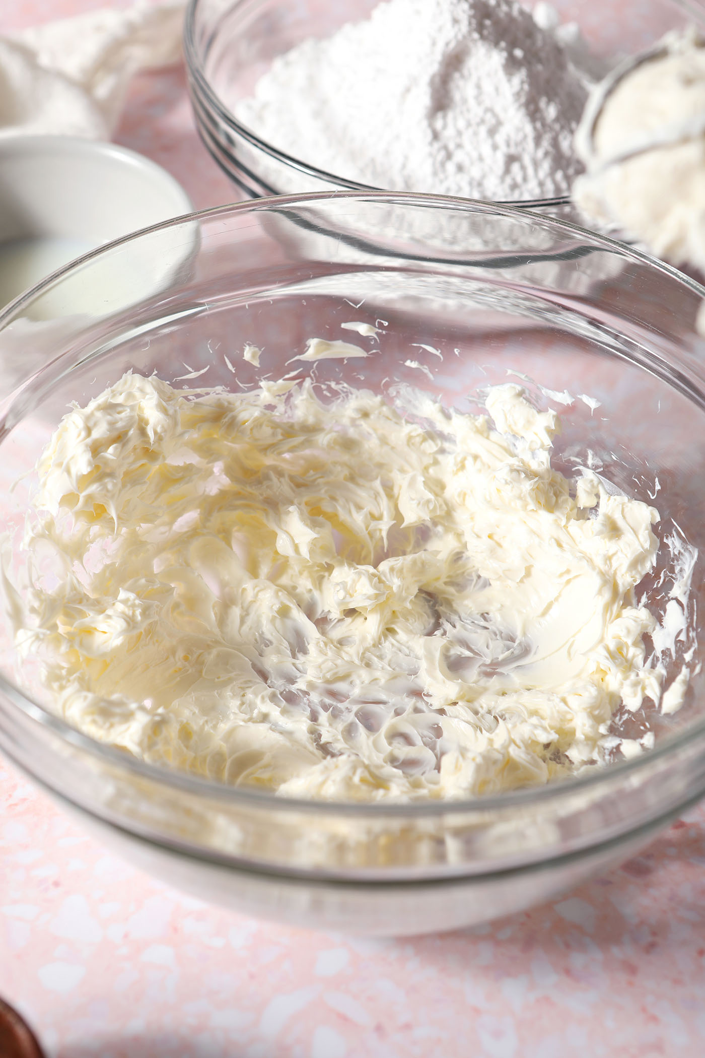 Cream cheese and butter in a bowl in a glass bowl on a pink surface next to other icing ingredients