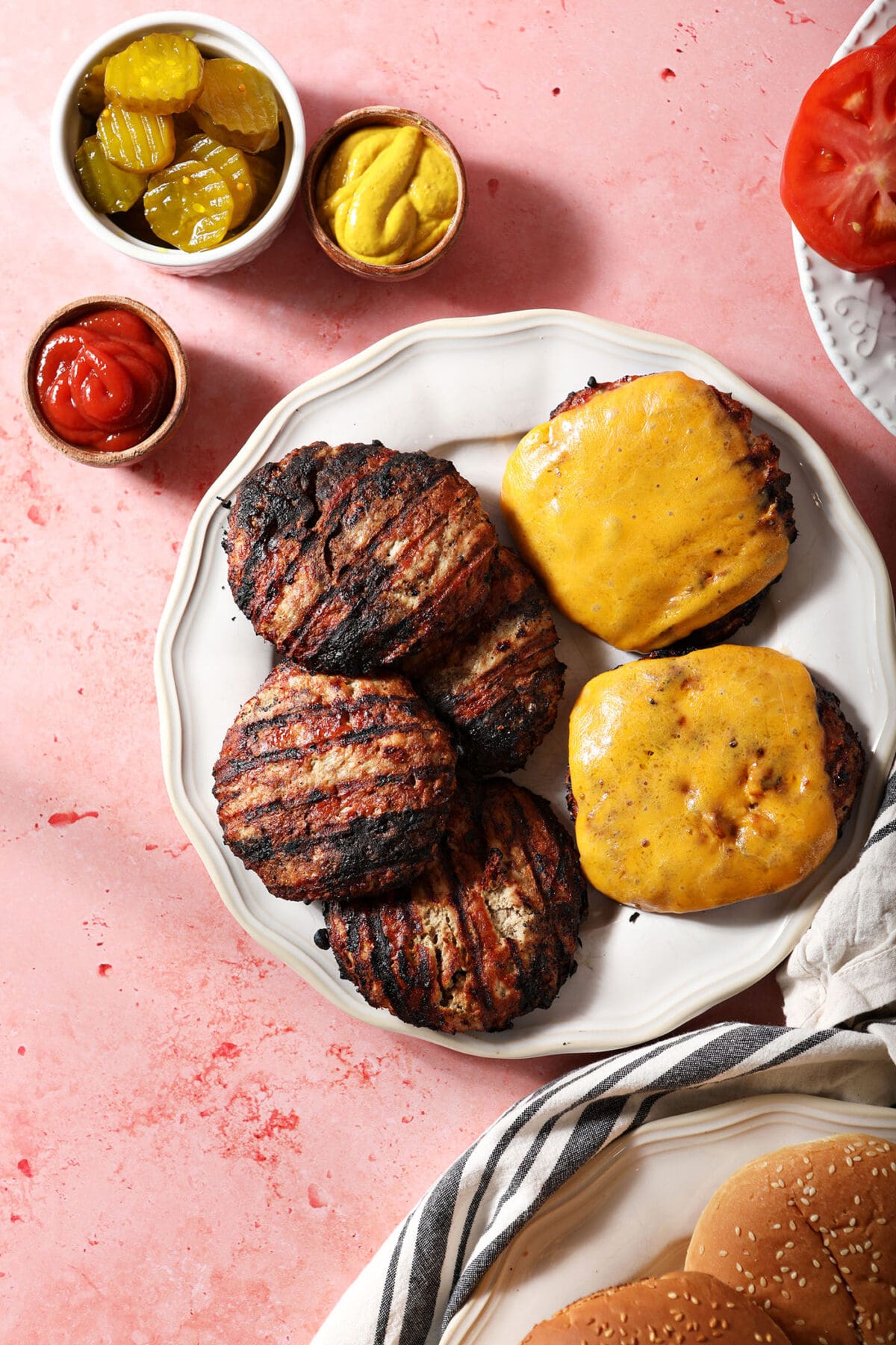 Grilled turkey burgers on a white plate next to bowls of condiments on a pink surface