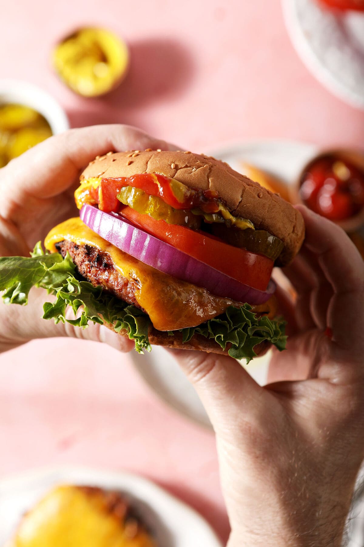 A person holds a turkey burger topped with cheese, lettuce, onion, tomato and more above a pink surface