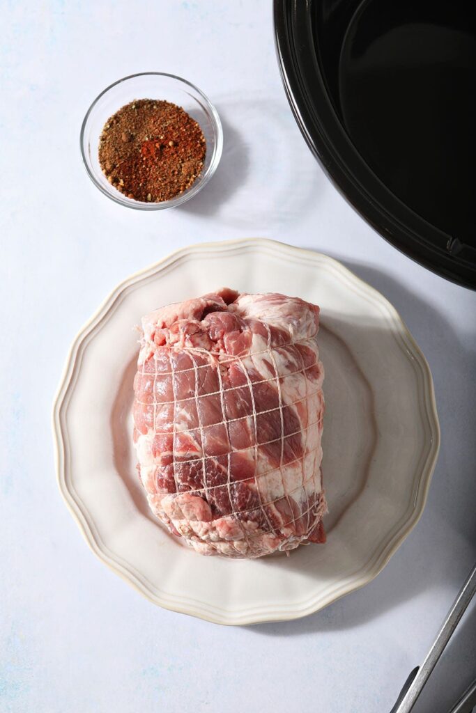 A raw pork shoulder on a plate next to a slow cooker and a bowl of seasoning