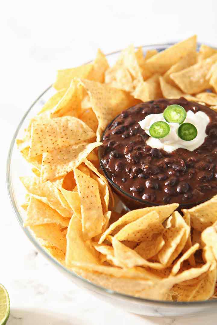 Black bean dip, garnished with sour cream and jalapeno slices, is shown in a bowl, surrounded by chips