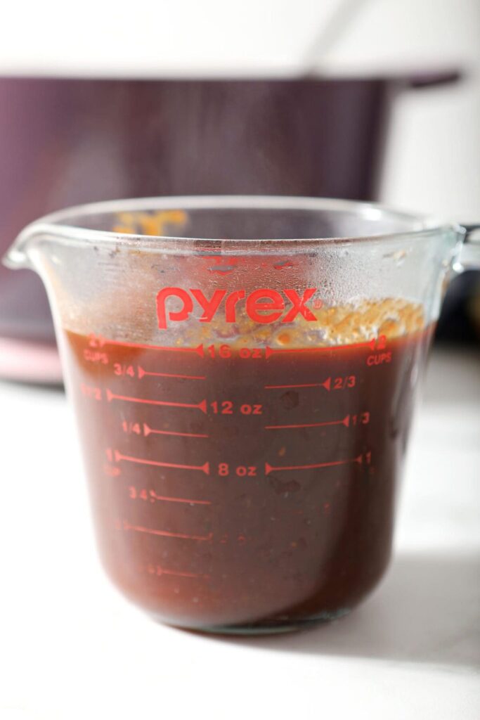 A glass measuring cup holds a serving of homemade barbecue sauce next to a purple Dutch oven