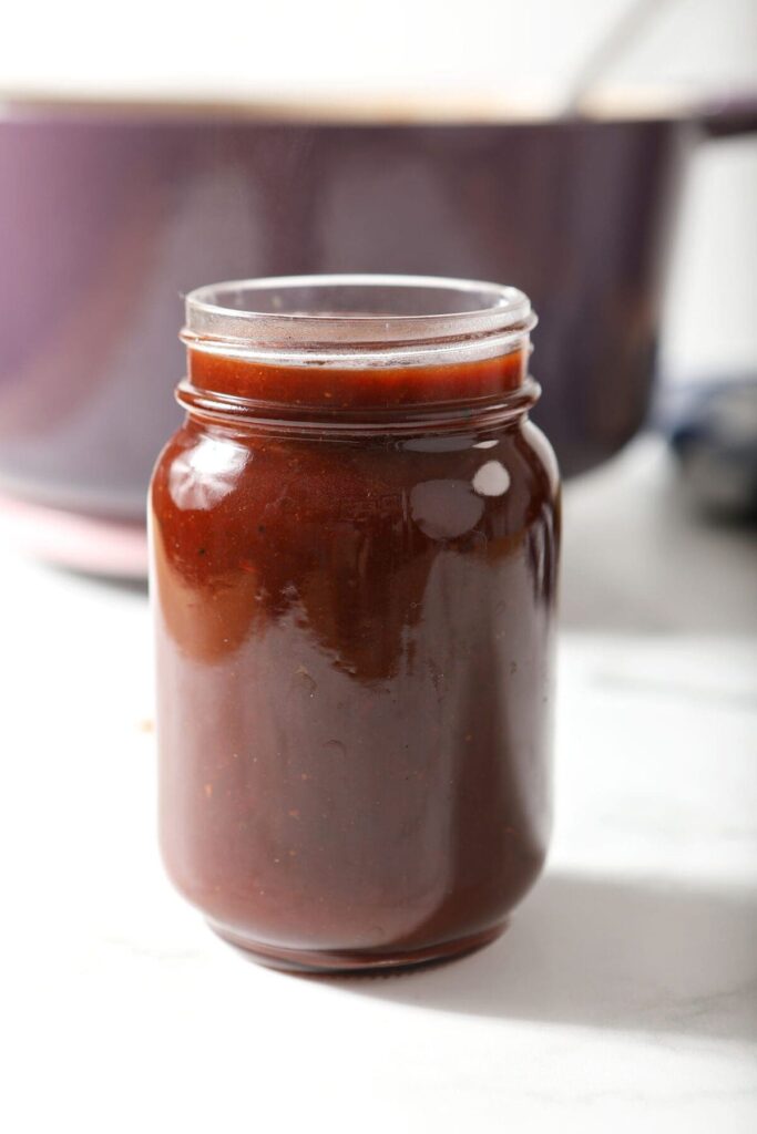 A jar of homemade BBQ sauce in front of a purple Dutch oven