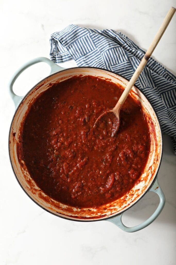 A pot of easy marinara sauce after simmering next to a blue checked towel with a wooden spoon