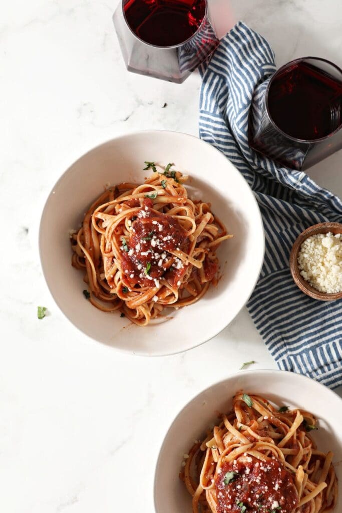 Two bowls of pasta covered in a simple marinara sauce on marble next to two glasses of red wine and a container of grated parmesan cheese