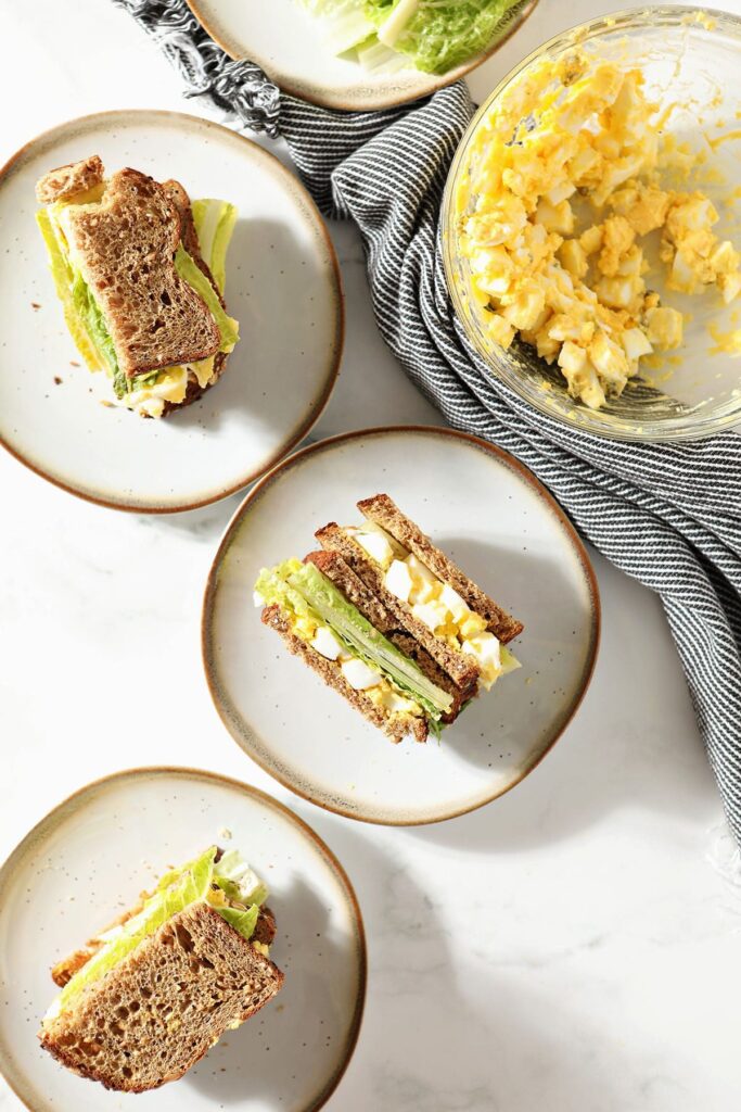 Three plates holding egg salad sandwiches next to a bowl of egg salad