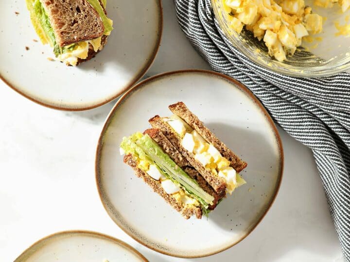 Three plates holding egg salad sandwiches next to a bowl of egg salad