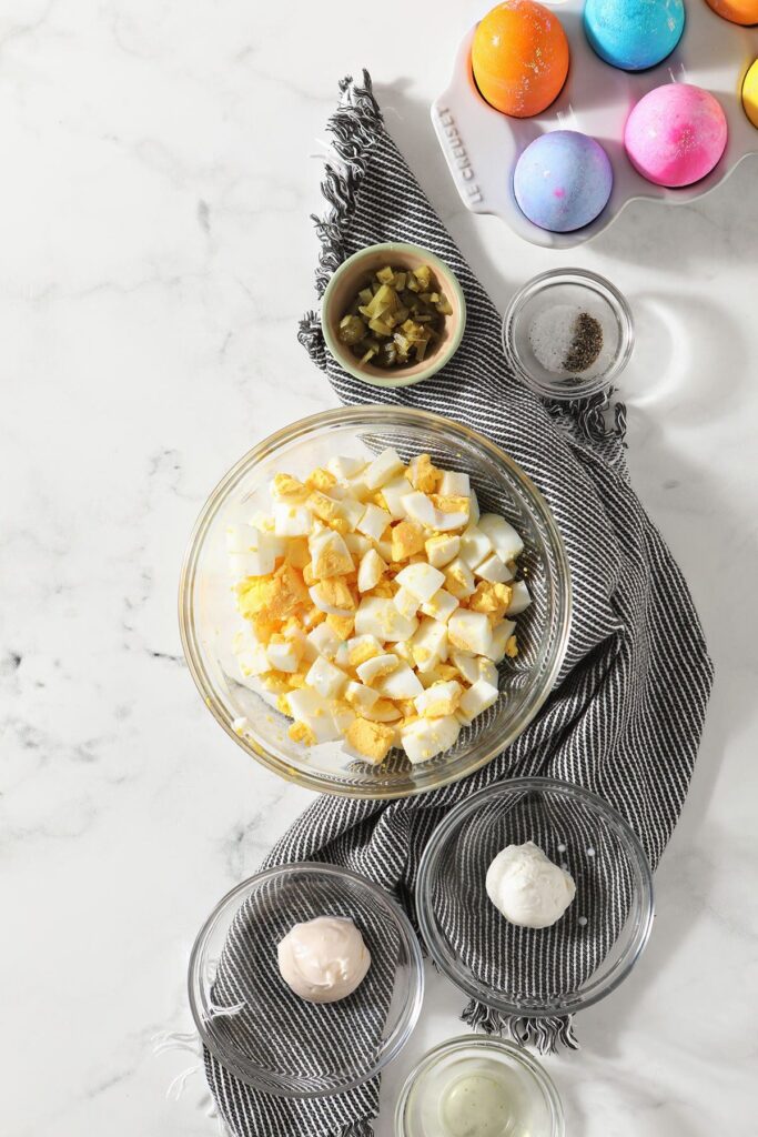 A bowl of hardboiled eggs with other bowls of ingredients for egg salad