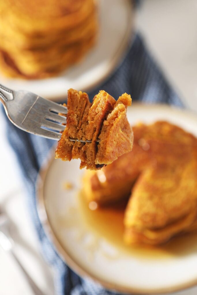 A fork holds a triangular bite of pancakes with a white plate of pancakes behind it