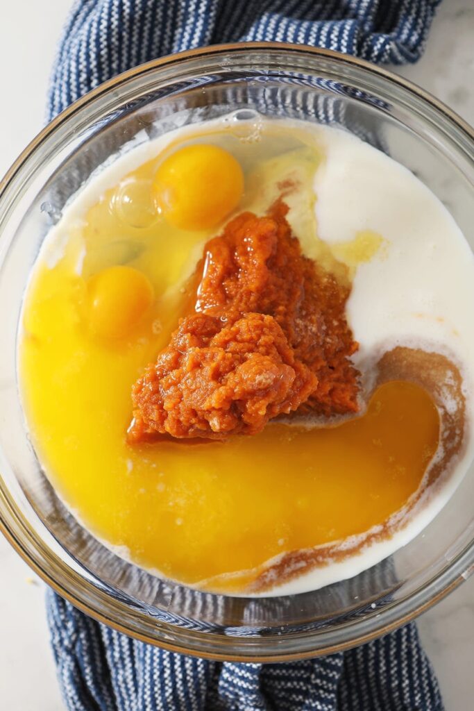 Pumpkin puree, buttermilk, eggs and other wet ingredients in a glass bowl on top of a blue towel