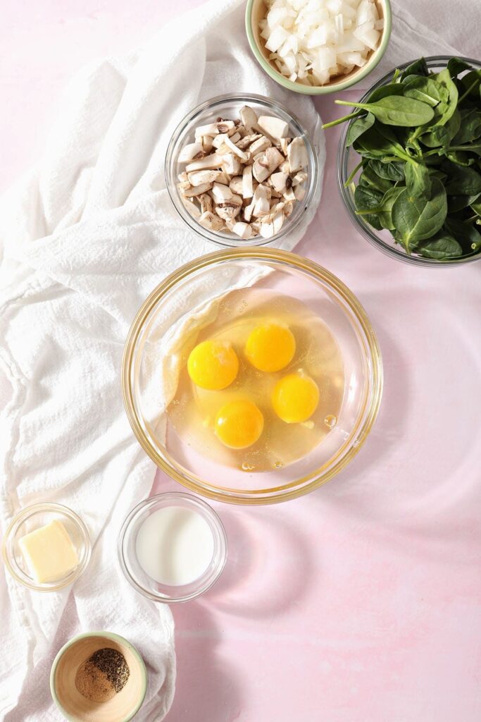 Ingredients to make veggie scrambled eggs on a pink countertop next to a white towel