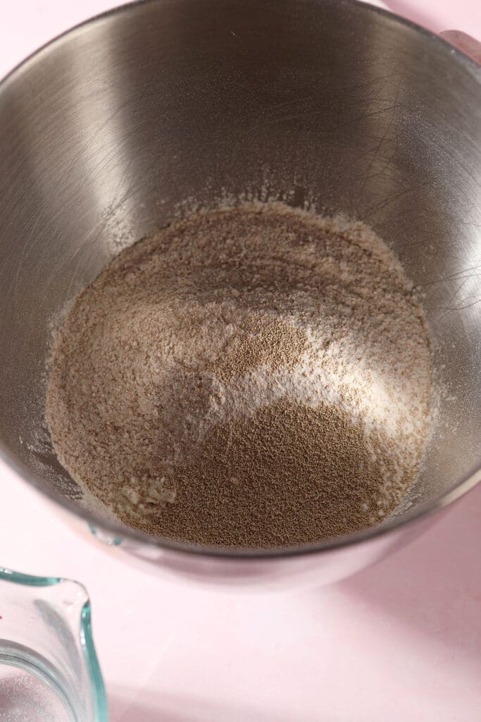 Flours and yeast in the bowl of a stand mixer on a pink surface