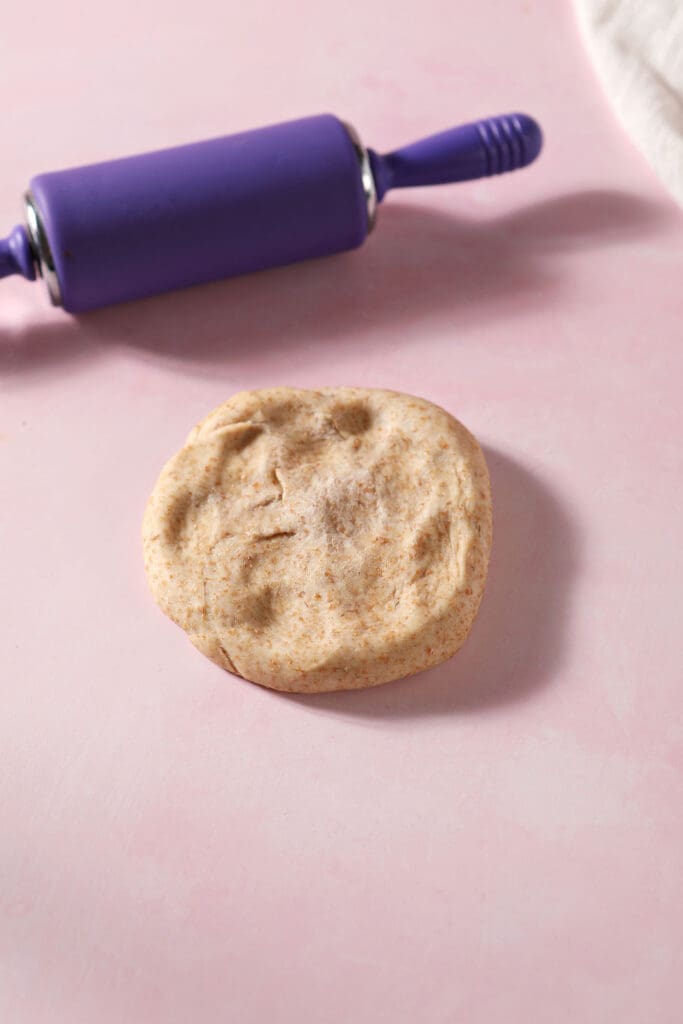 A round of pizza dough pressed down on a pink surface next to a purple rolling pin