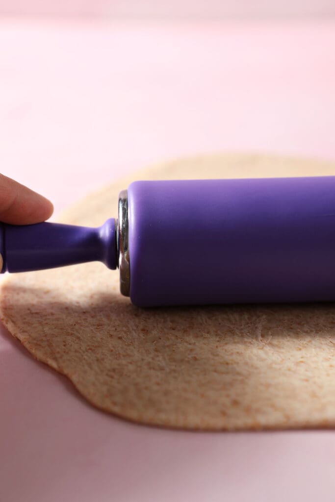 A purple rolling pin rolls out pizza dough on a pink surface