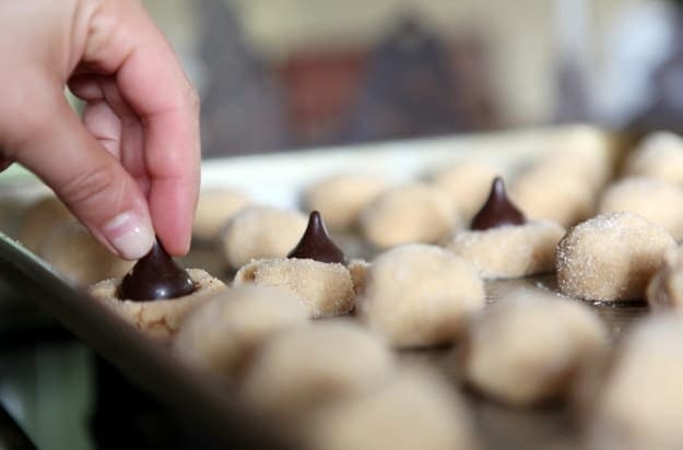 A hand presses a dark chocolate Hershey's Kiss into a peanut butter cookie on a baking sheet