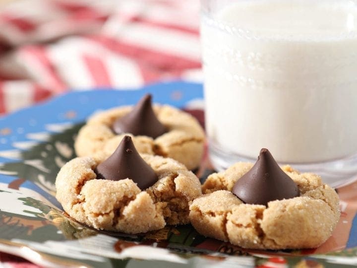 Three Dark Chocolate Peanut Blossoms sit on a blue painted plate next to a glass of milk on top of a red and white striped towel