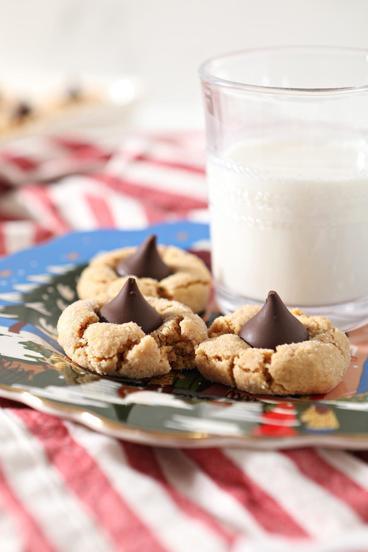 Three Dark Chocolate Peanut Blossoms sit on a blue painted plate next to a glass of milk on top of a red and white striped towel