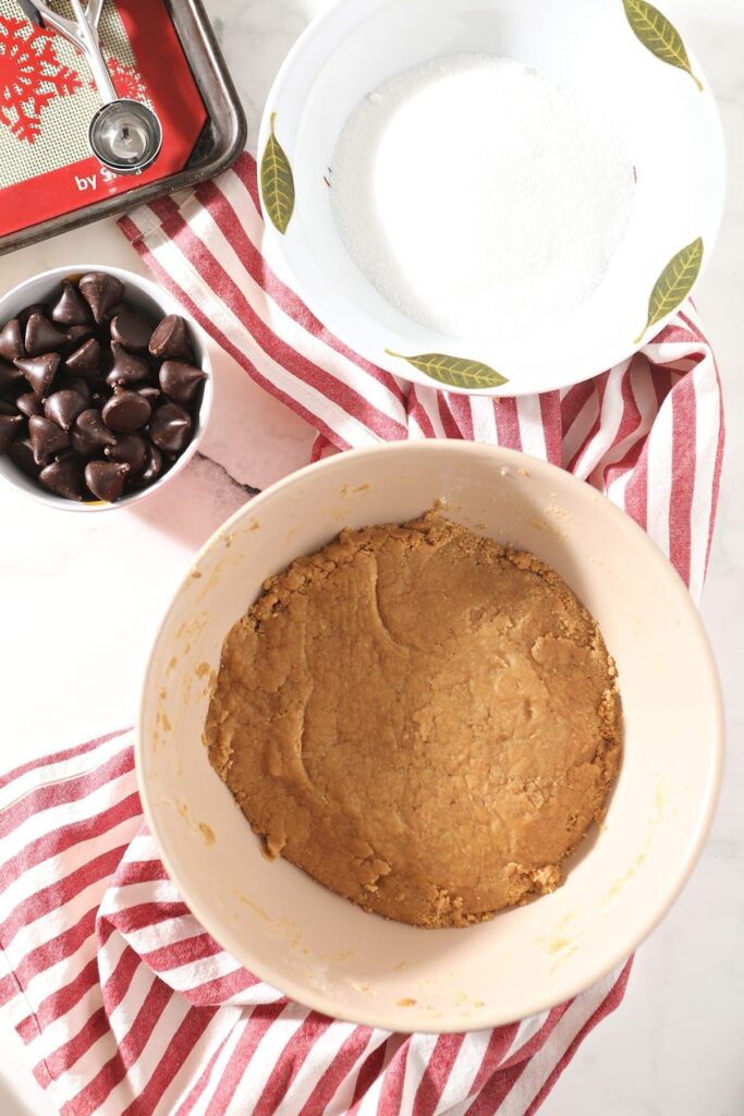 A large mixing bowl holds peanut butter cookie batter and sits next to a bowl of Dark Chocolate Hershey's Kisses and another one of granulated sugar