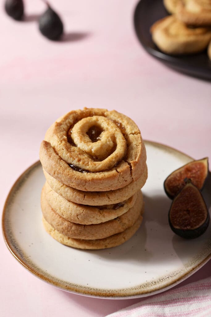 a stack of pinwheel cookies on a white plate next to sliced figs on a pink surface