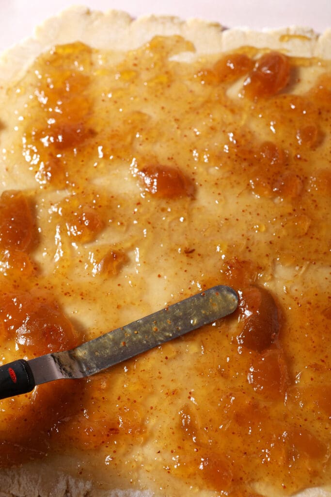 an offset spatula spreads fig preserves onto rolled sugar cookie dough