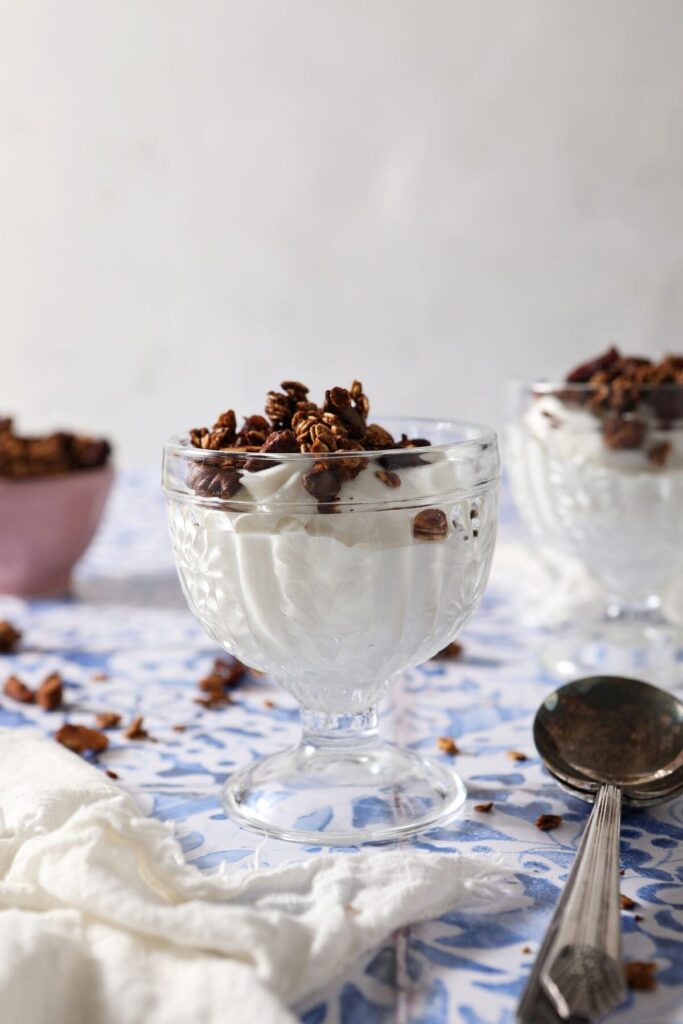 Two glass containers of yogurt topped with granola on a blue tiled surface with spoons