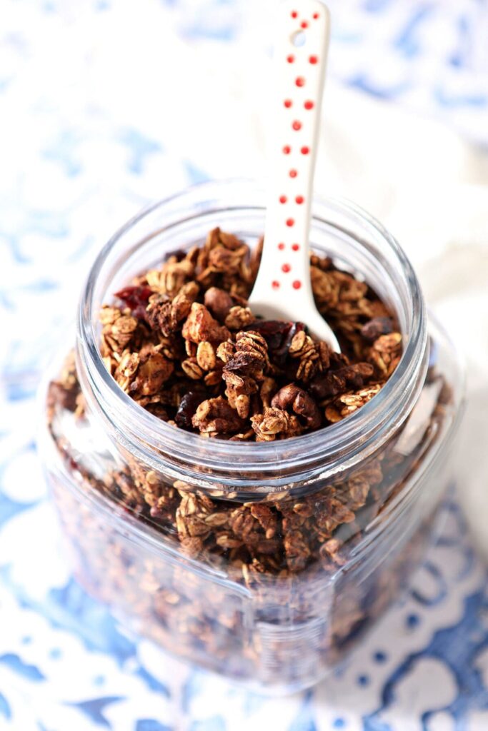 A jar of homemade maple nut granola with a spoon in it on a blue tile surface