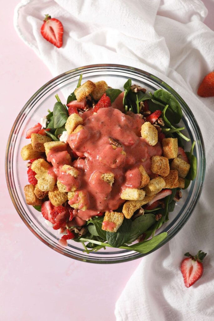 Strawberry salad dressing on top of a spinach salad in a bowl before tossing on a pink surface