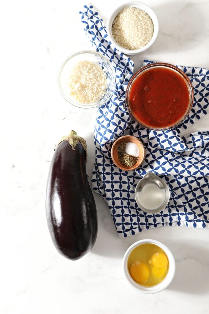 Ingredients to make baked eggplant parmesan on marble with a patterned blue and white towel