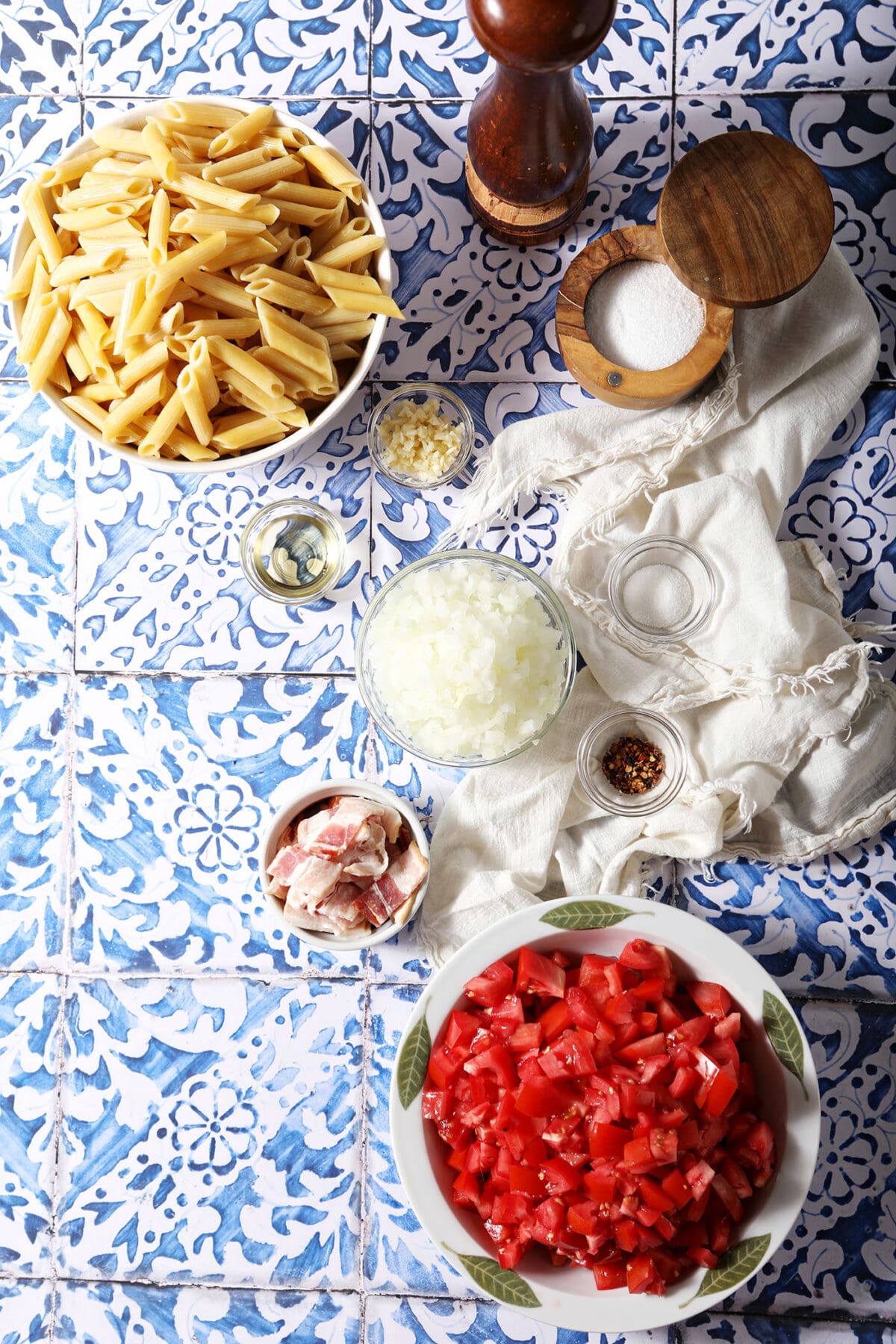 Ingredients to make Penne Arrabbiata in bowls on a blue and white tiled background with white linens