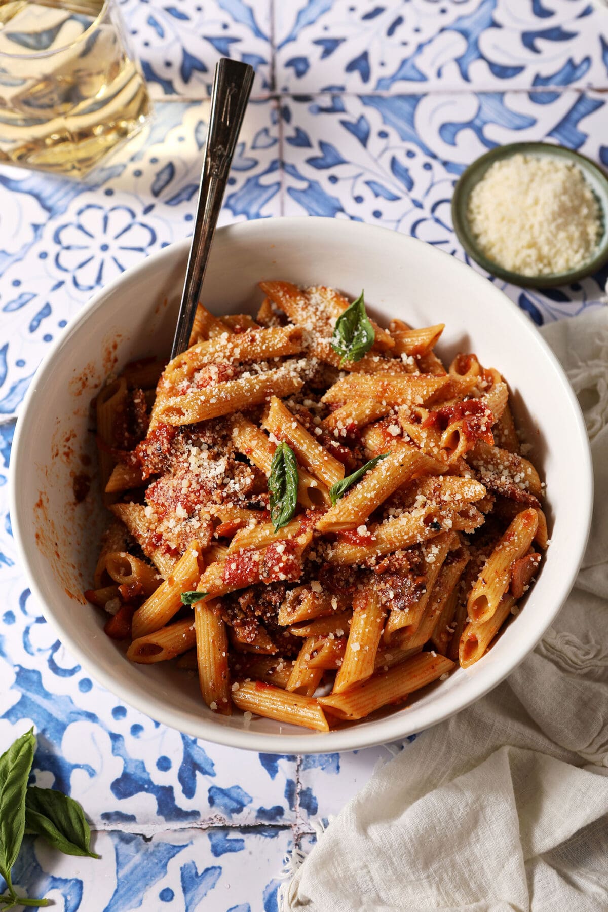 A white bowl holds a serving of Penne Arrabbiata garnished with fresh basil and parmesan on a blue and white tiled surface