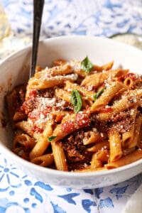 Side angle of a white bowl holding Penne Arrabbiata garnished with fresh basil and parmesan on a blue and white tiled surface