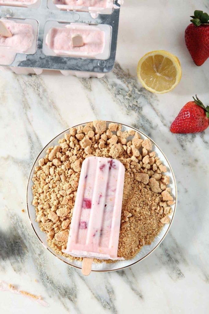 A Roasted Strawberry Cheesecake Popsicle sits on a plate of Graham cracker crumbs before consuming