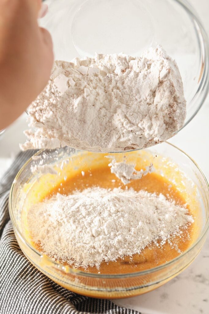 Flour is poured on top of orange batter in a glass bowl