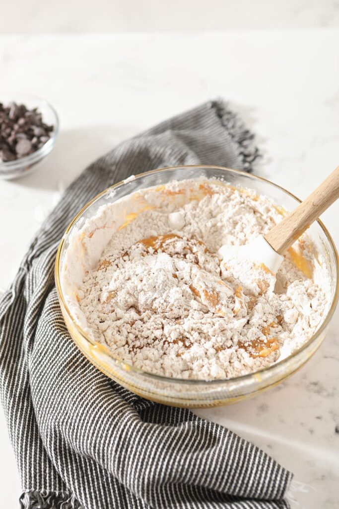 Flour is mixed into orange batter with a white spatula in a glass bowl on top of a gray striped towel