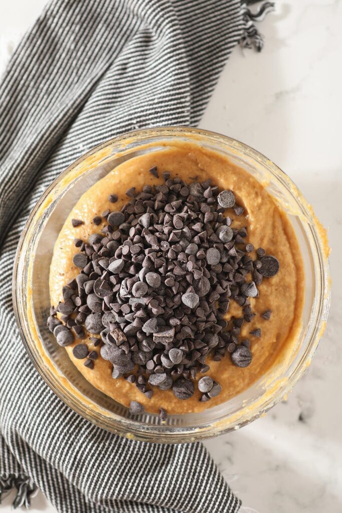 Chocolate chips on top of pumpkin bread batter in a glass bowl on top of a gray striped towel