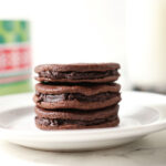 Three chocolate sandwich cookies with a ganache filling stacked on top of each other on a white plate in front of a Christmas cookie tin on marble