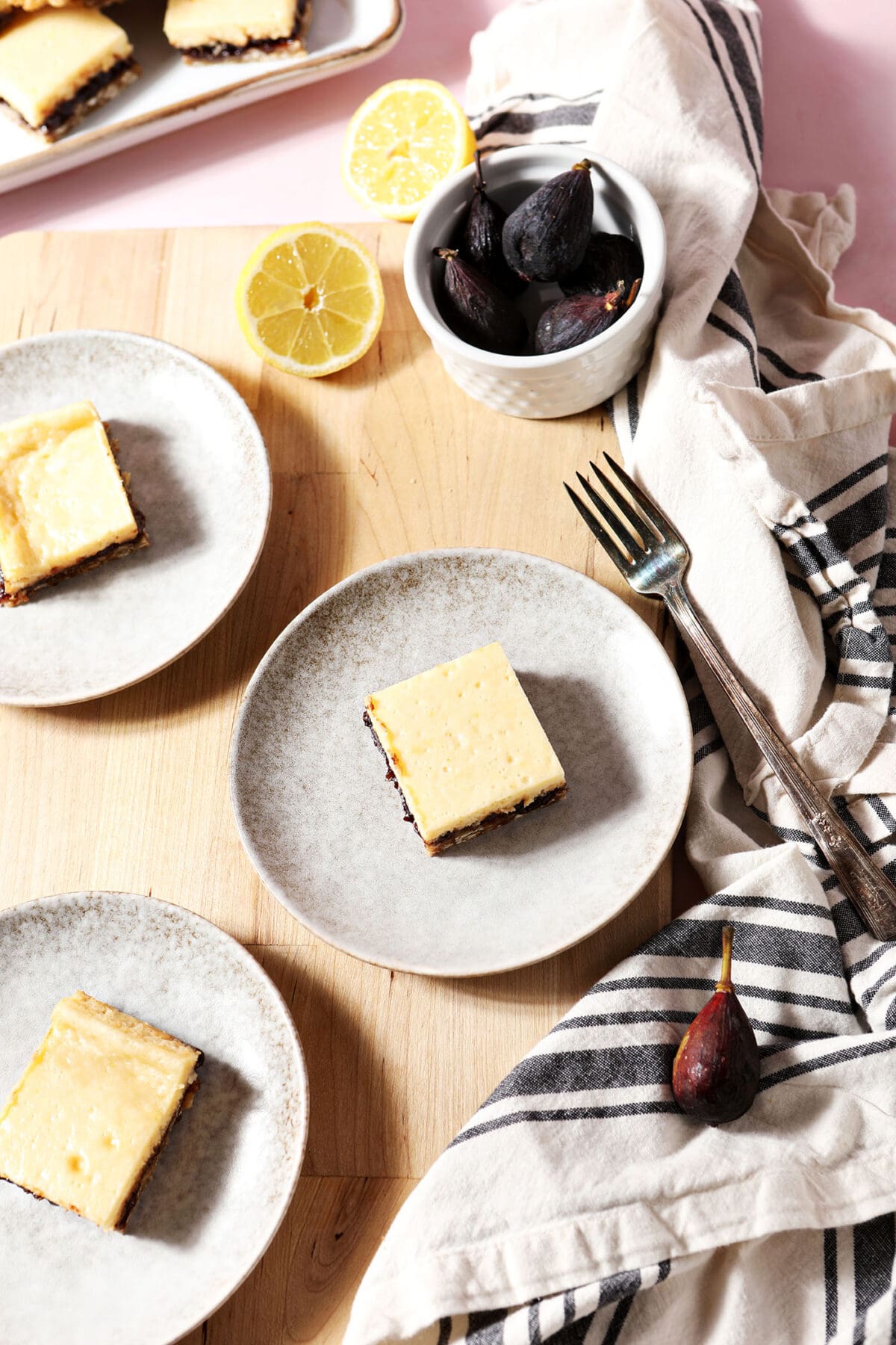 three lemon cheesecake bars with fig on white plates with figs and lemons and a striped linen on a wooden surface with pink