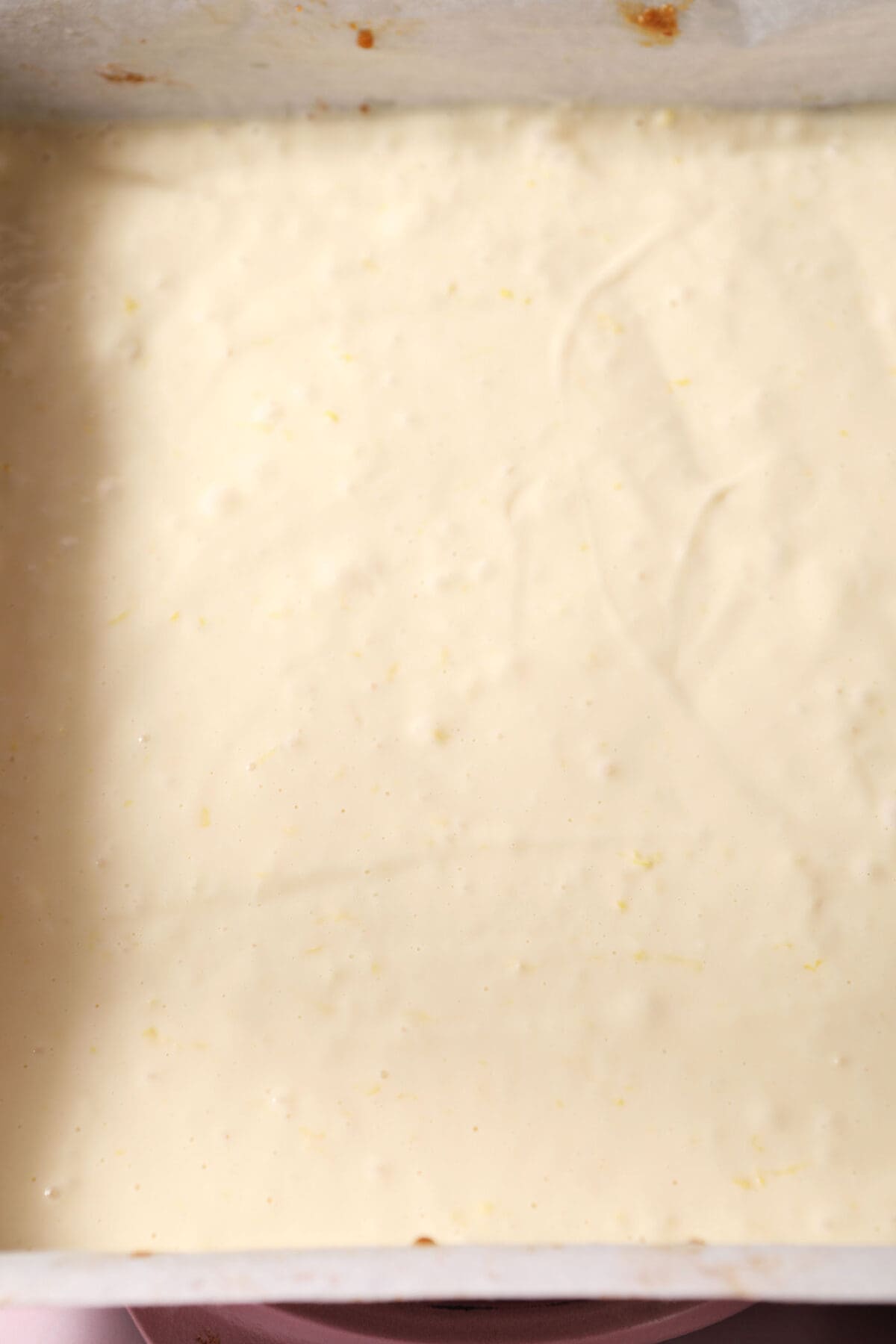 cheesecake filling on top of a crust before baking in a square baking pan on a pink surface