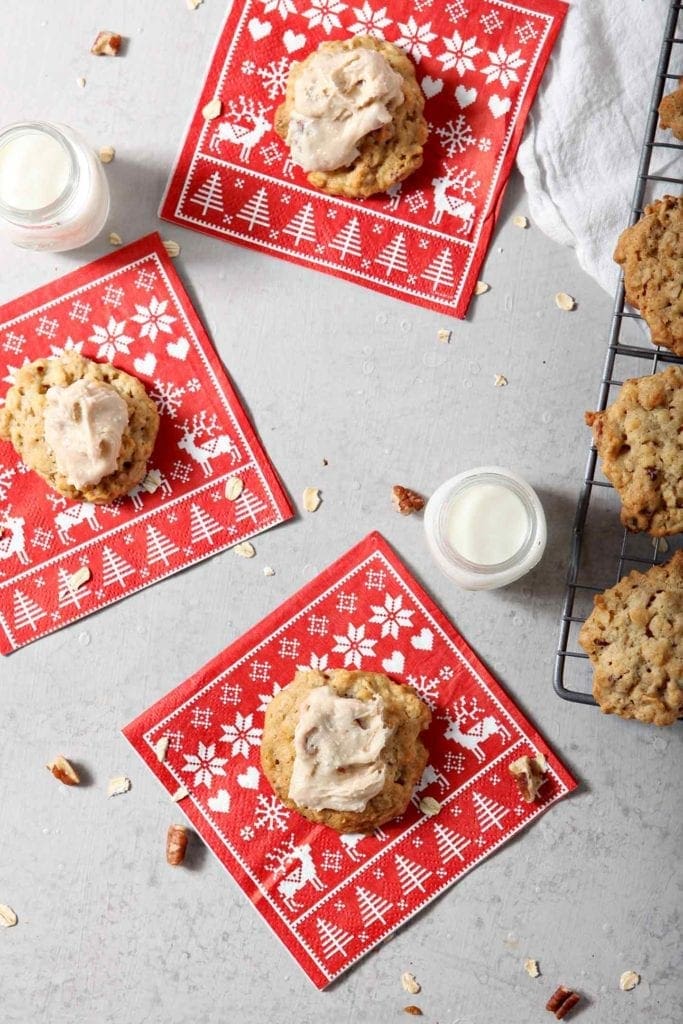 Three iced Quebec Maple Pecan Drop Cookies sit on red patterned napkins with small glasses of milk next to the wire cooling rack with more cookies