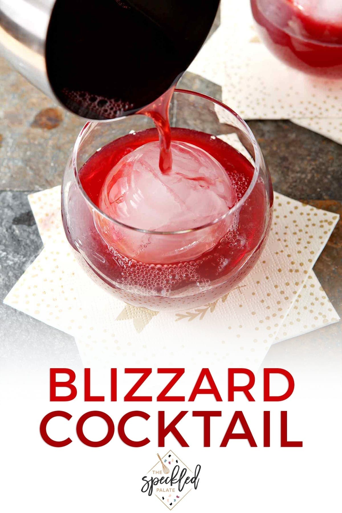 A silver cocktail shaker pours red liquid into an old fashioned glass with a large spherical ice cube with the text 'blizzard cocktail'