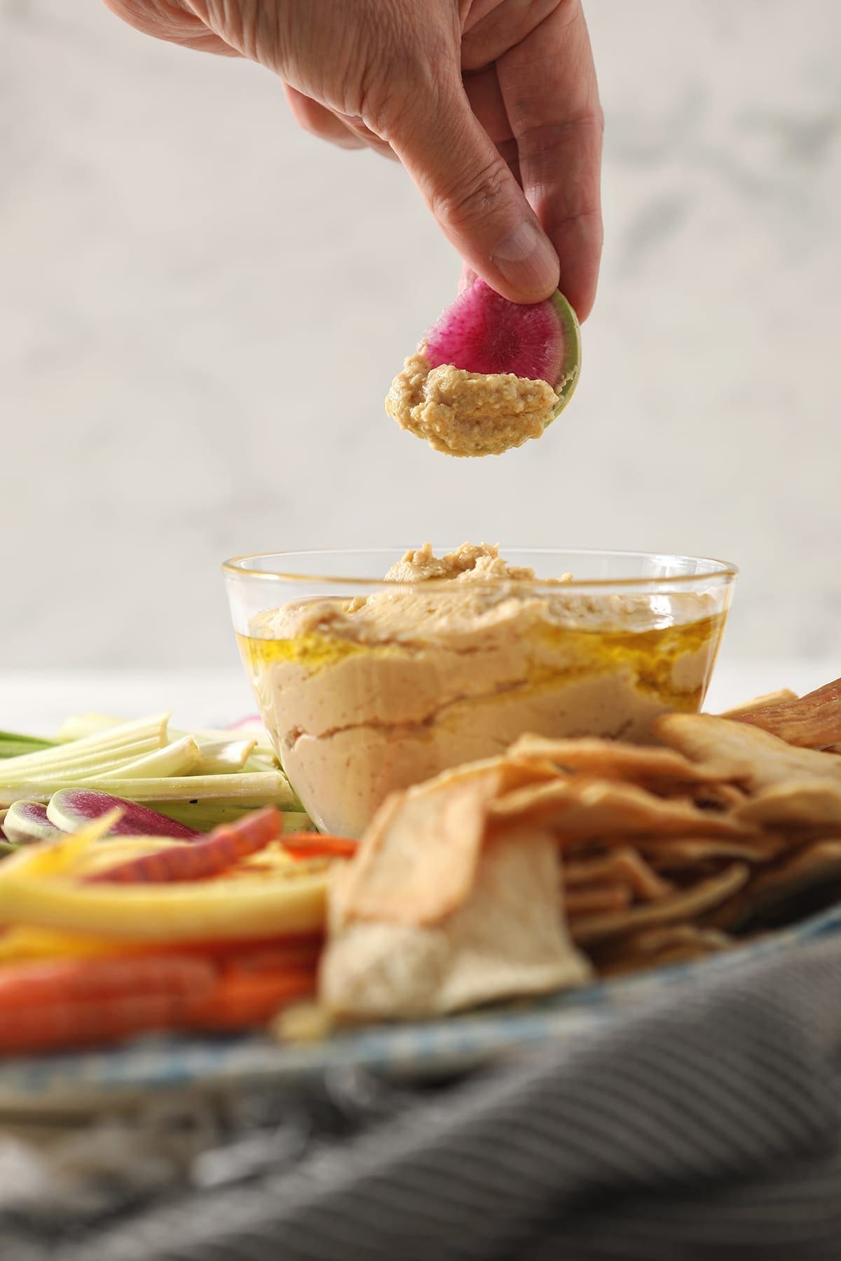 A woman dips a watermelon radish into a clear glass bowl of homemade garlic hummus