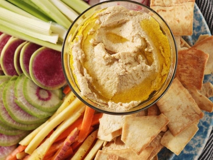 Hummus, drizzled with olive oil, sits in a glass container surrounded by radish, celery and carrot slices and pita chips