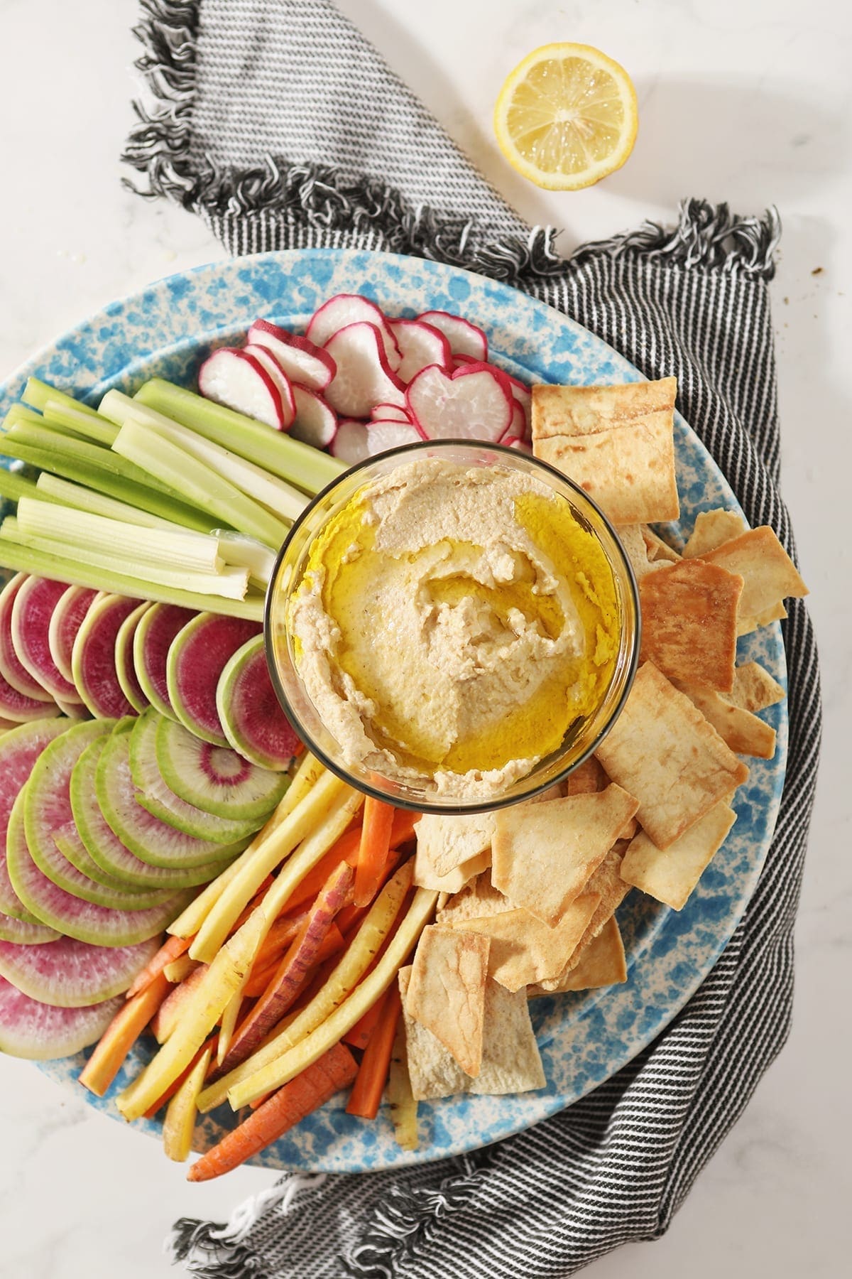 Garlic Hummus, drizzled with olive oil, sits in a glass container surrounded by radish, celery and carrot slices and pita chips