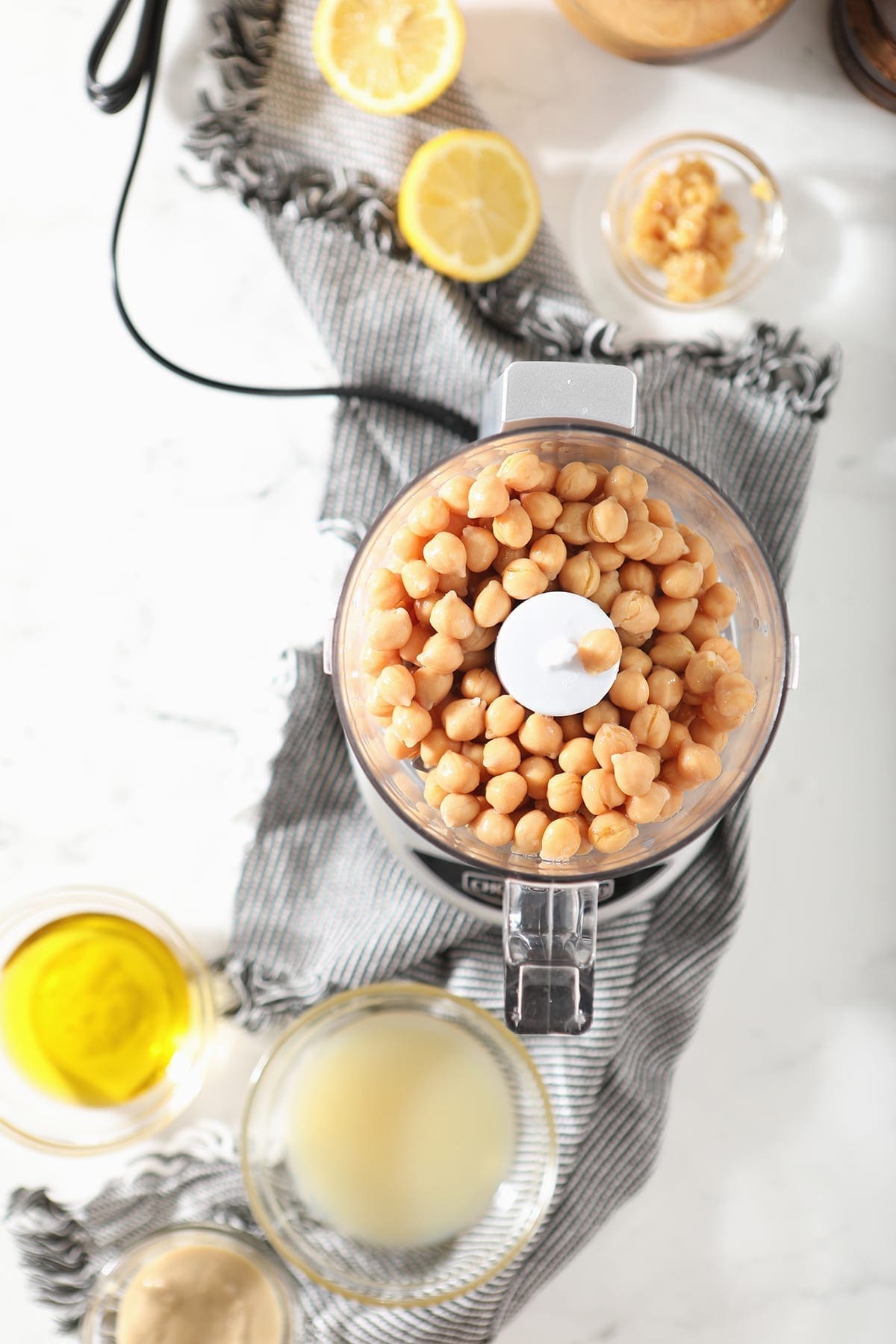 Chickpeas in a small food processor, surrounded by other ingredients on a marble surface