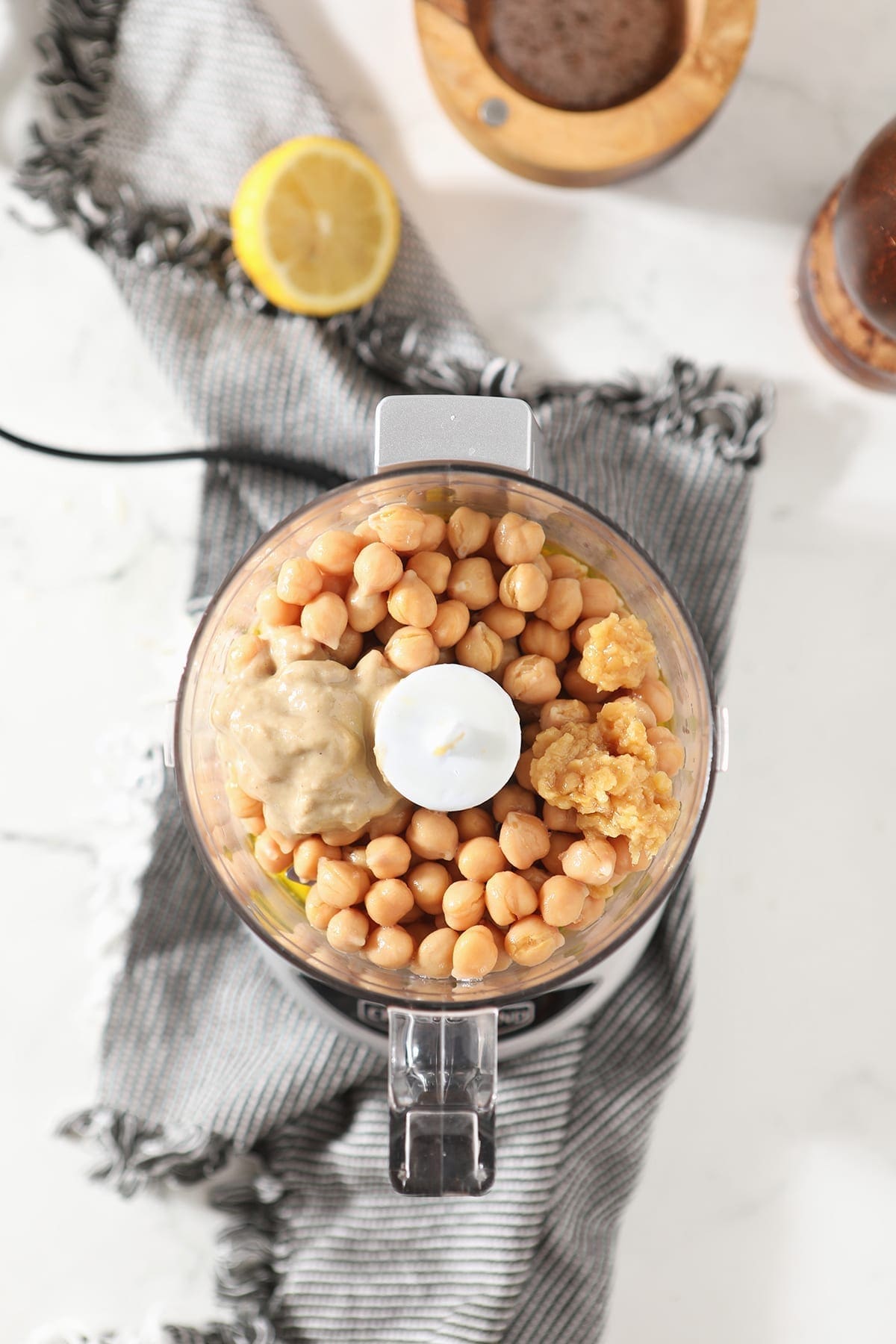 Tahini and garlic sit on top of whole chickpeas in a food processor on a striped towel on marble