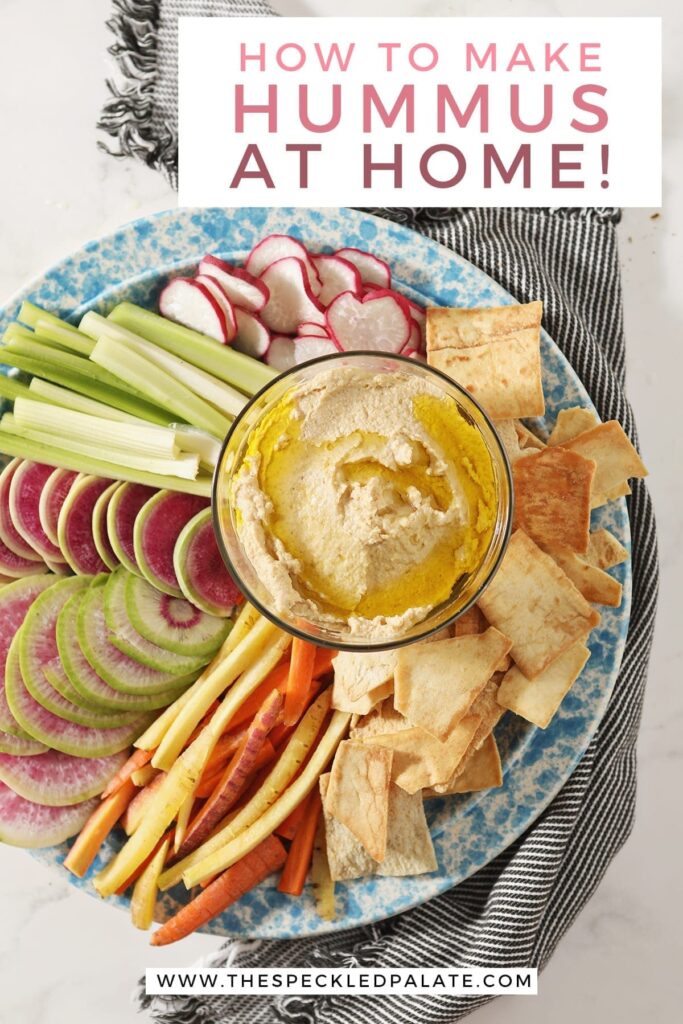 Hummus, drizzled with olive oil, sits in a glass container surrounded by radish, celery and carrot slices and pita chips with text "How to Make Hummus at Home!"