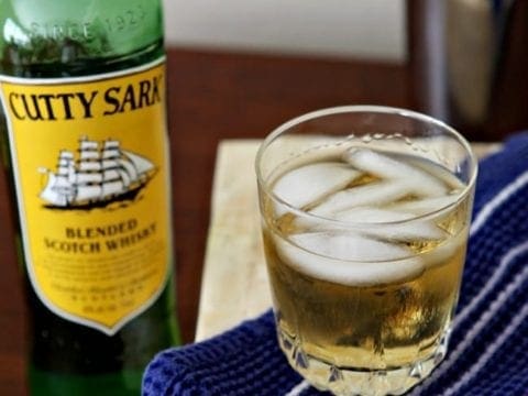 A glass of Cutty Sark on the rocks sits with a bottle of the scotch blend on a table with a red, white and blue towel