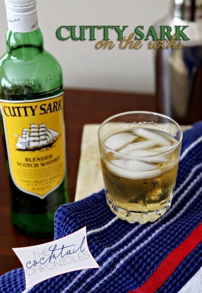 A glass of Cutty Sark on the rocks sits with a bottle of the scotch blend on a table with a red, white and blue towel
