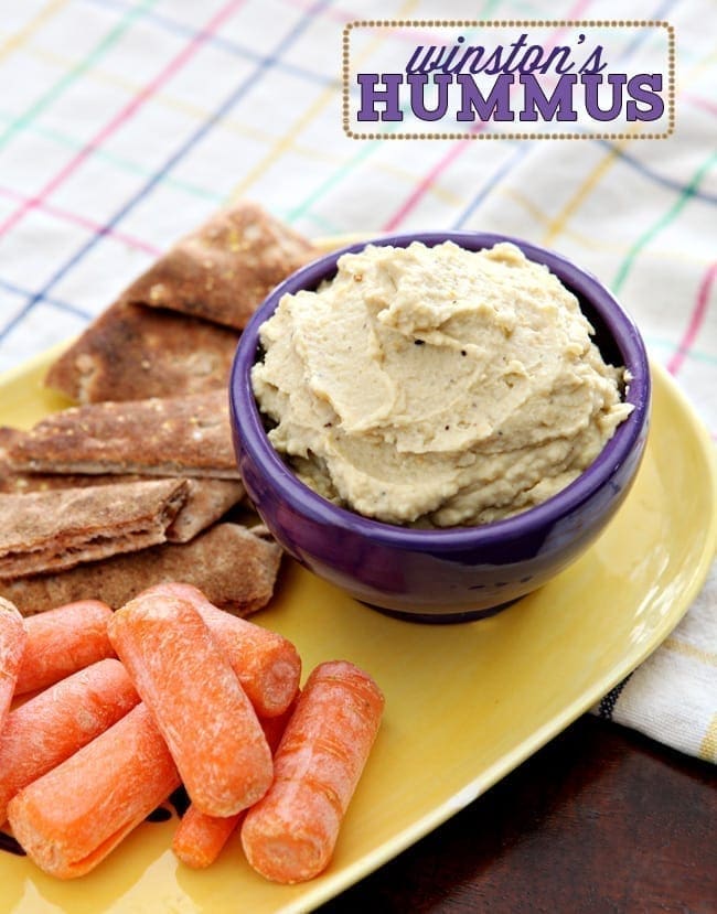 A purple bowl holds homemade Hummus and sits on a yellow plate with baby carrots and pita strips