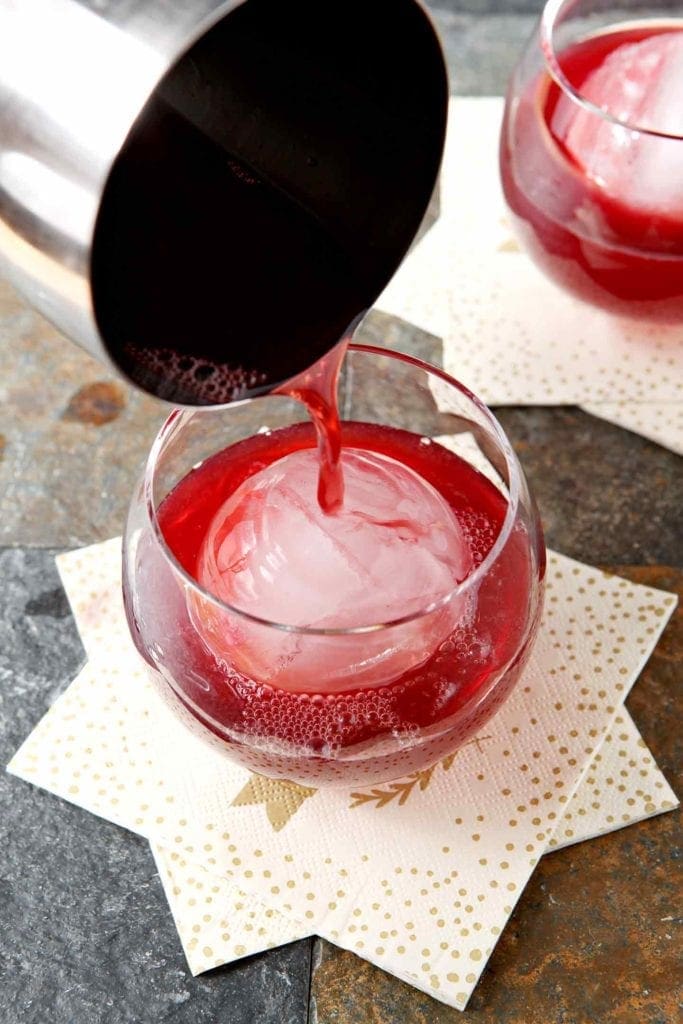 A cranberry cocktail is poured into a rocks glass with a giant spherical ice cube